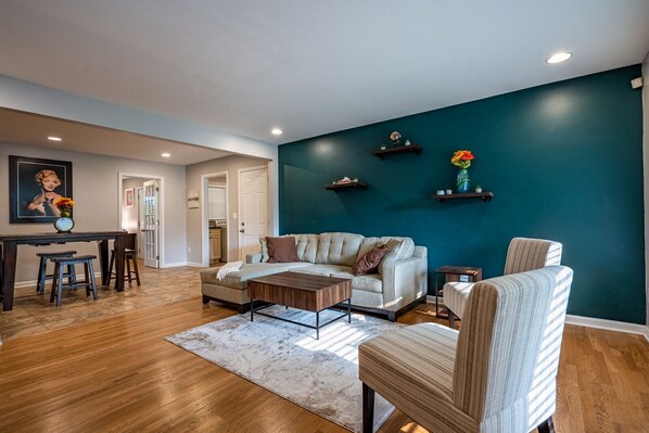 A living room with a sofa, coffee table, and chairs, a dining area with a table and chairs, and a kitchen in the background. - Discover Your Beautiful Dream Vacation Rental at The Battery - Truist Park!  (Smyrna)