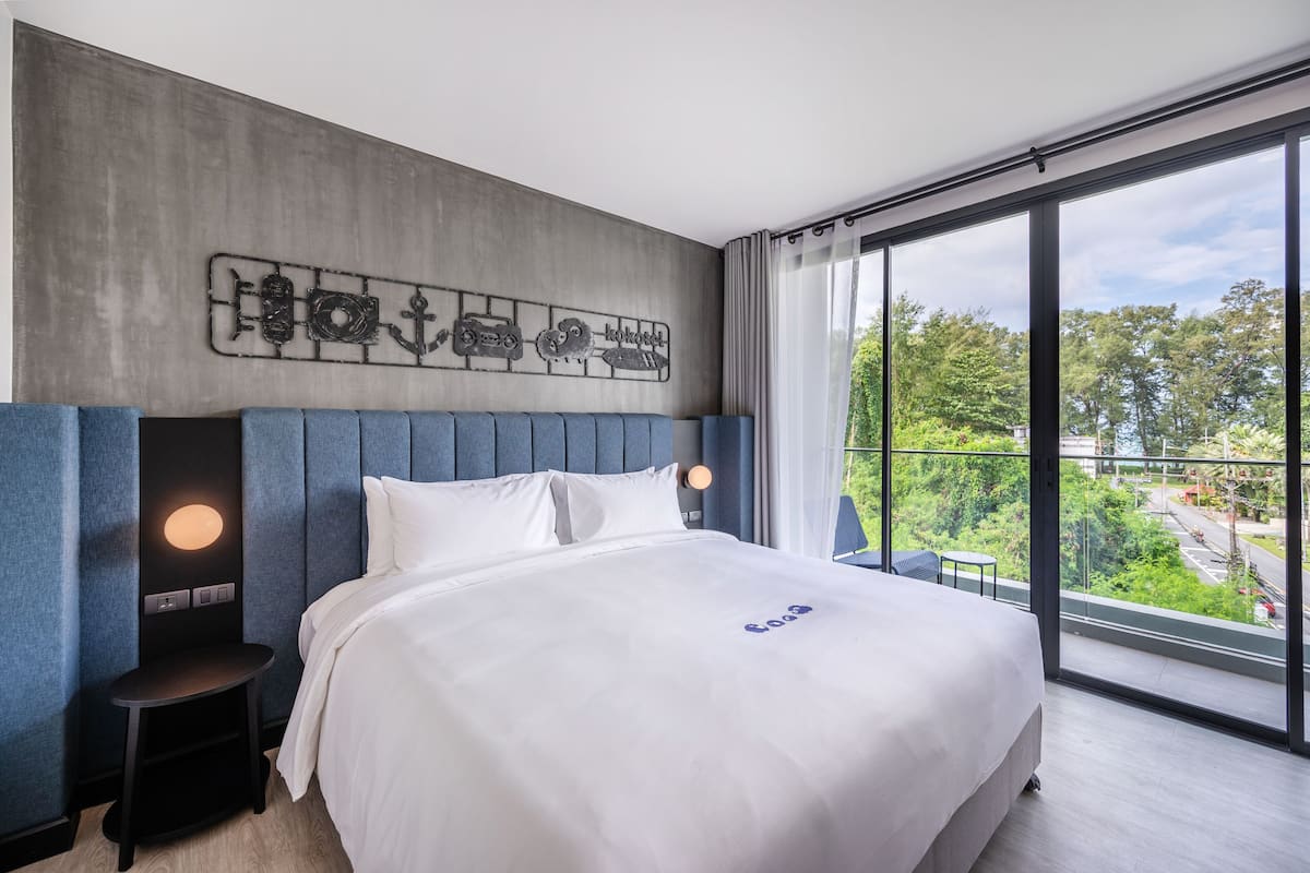 A modern hotel room with a large bed, a headboard with a cityscape design, a bedside table with a lamp, and a large window offering a view of the city.