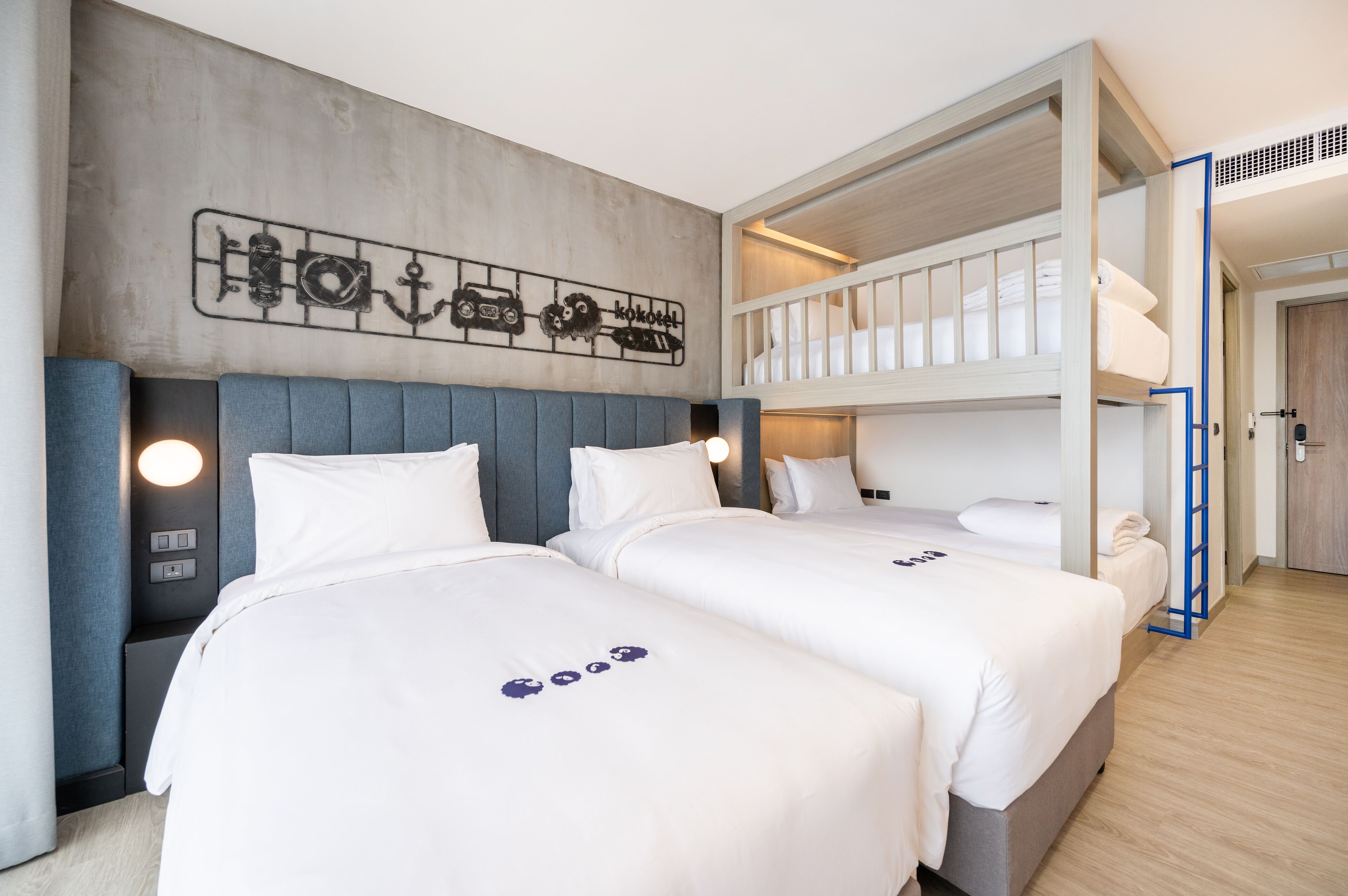 A hotel room with two beds, a bunk bed, a concrete wall with a car-themed decoration, and a wooden floor.