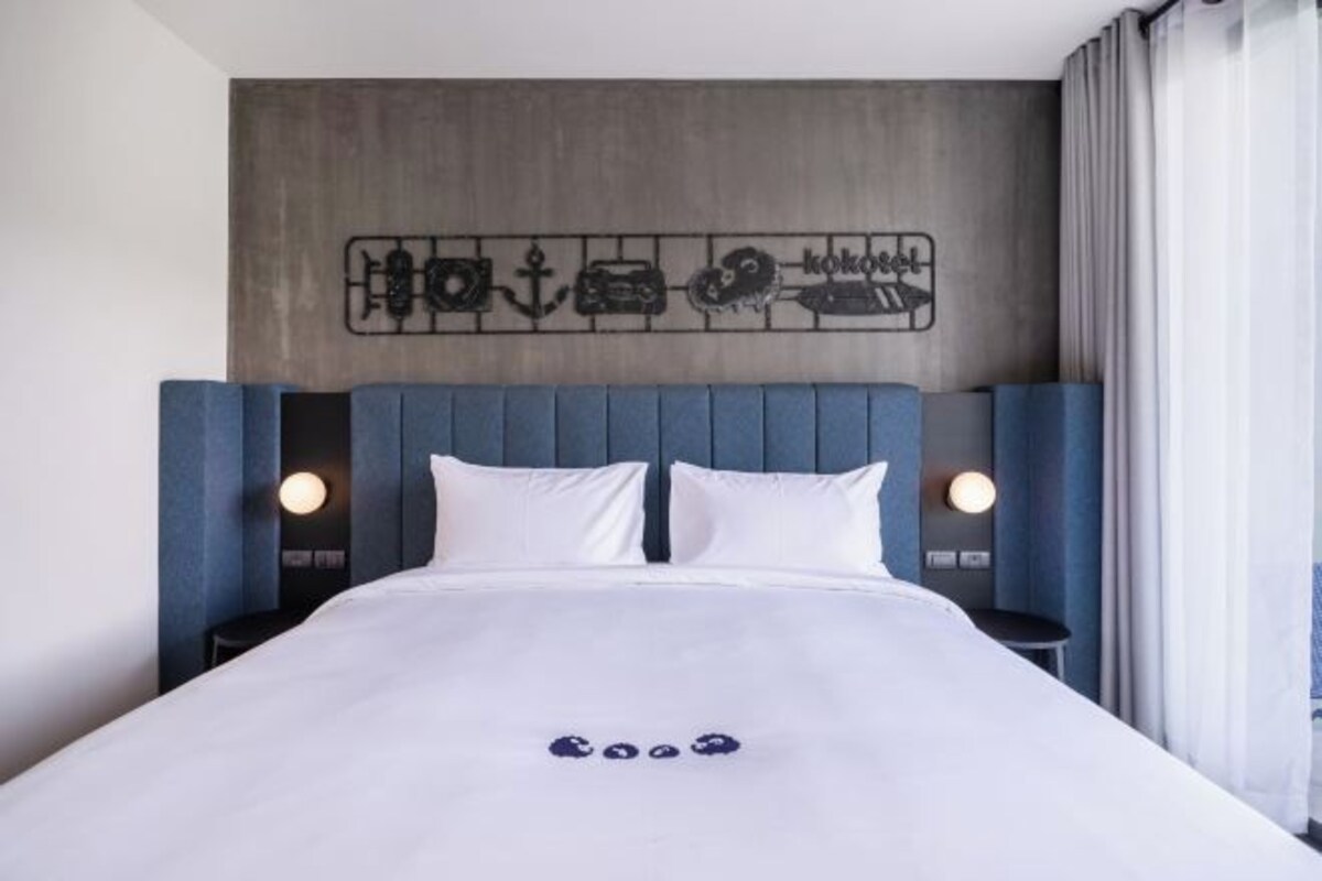 Una habitación de hotel moderna con una cama grande, un cabecero con panel decorativo, dos mesitas de noche con lámparas y una ventana con cortinas.