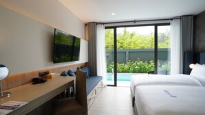 Habitación de hotel con cama, escritorio, silla, televisor y puerta corrediza de vidrio a un balcón con vegetación.