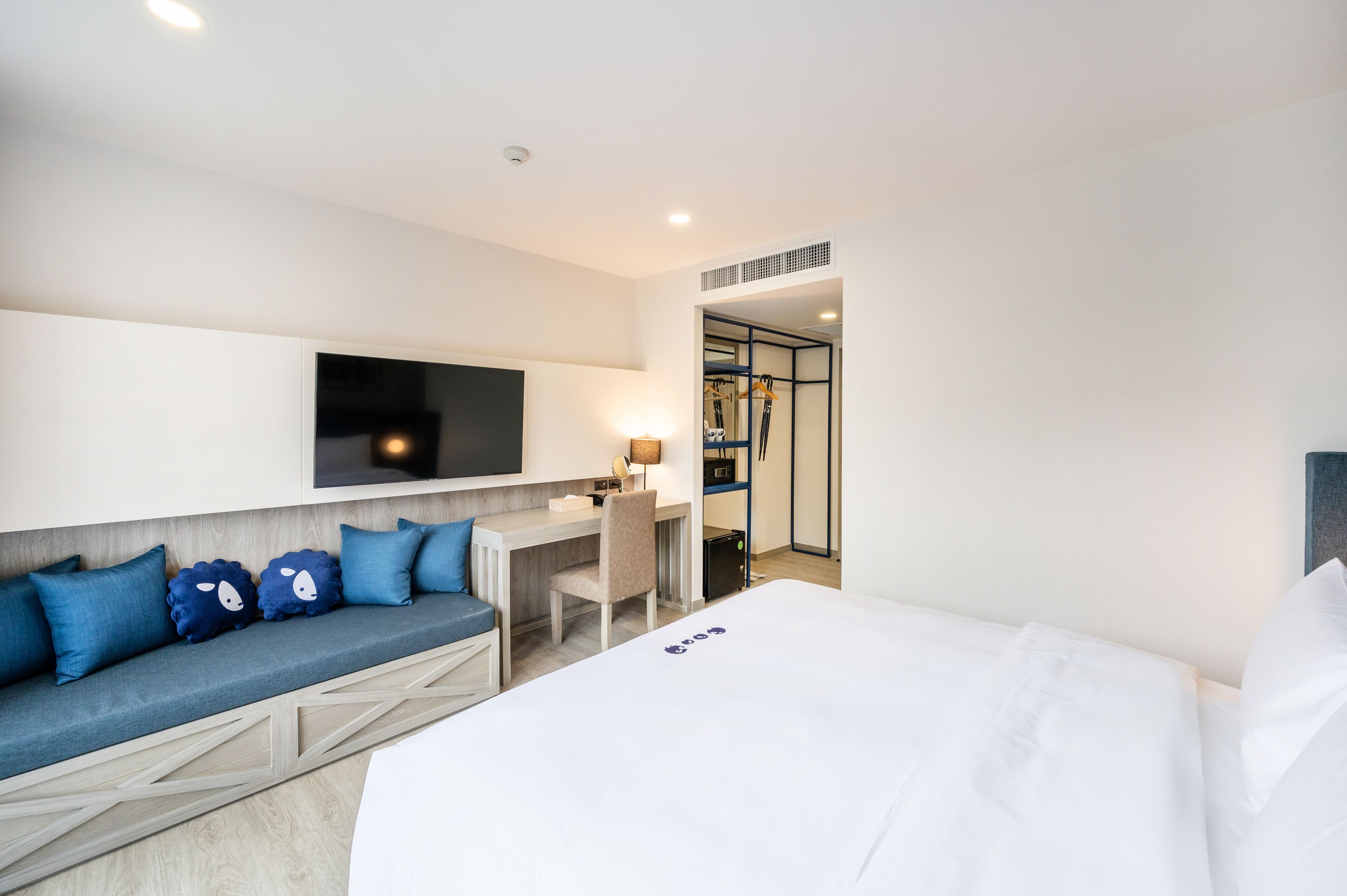 Habitación de hotel con cama, sofá con cojines azules, escritorio con silla, televisor montado en la pared y vestidor.