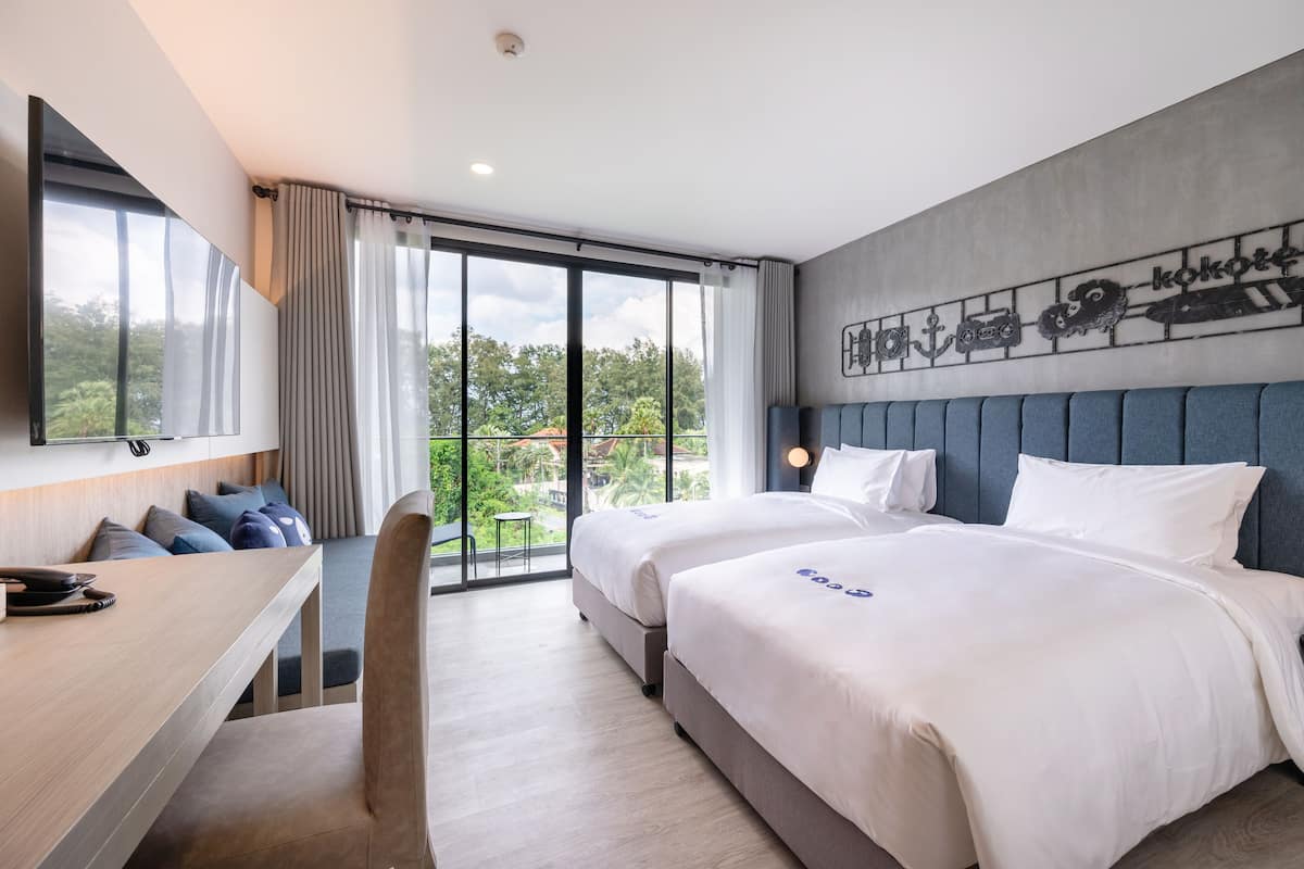 A modern hotel room with two beds, a desk, and a large window with a view of greenery.