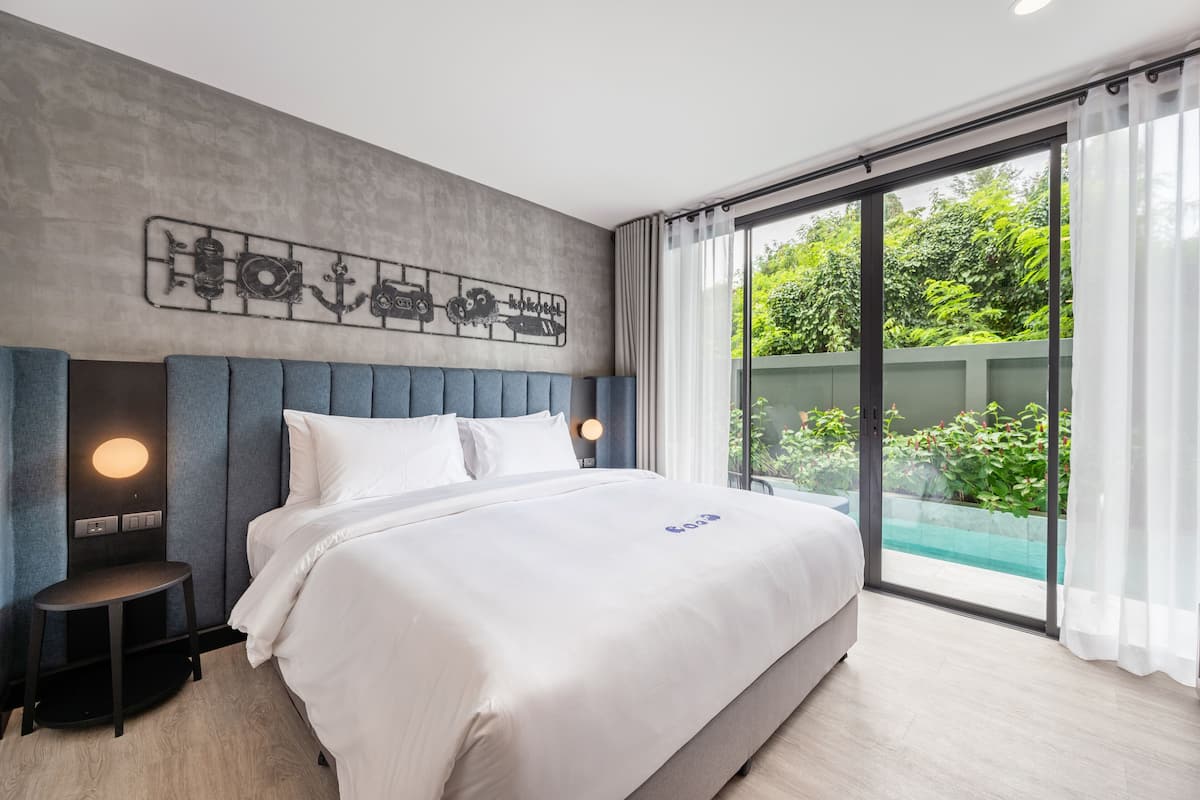 A modern hotel room with a large bed, a headboard with a light fixture, a small side table, and a view of greenery through a sliding glass door.