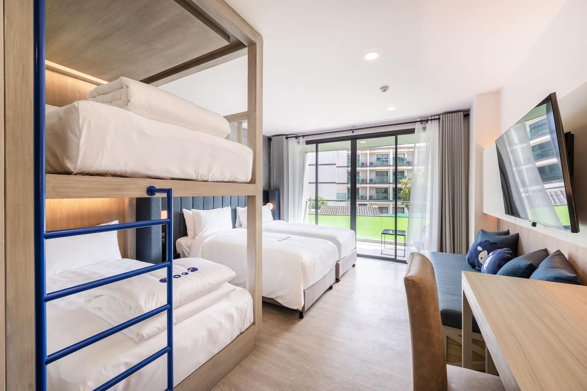 A hotel room with bunk beds, a flat-screen TV, a dining table, and a view of a building and greenery outside.