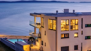 A modern two-story house with a balcony overlooking a body of water, a swimming pool, and a well-lit interior. - Jimmys Beach Privlaka: Penthouse dream, right by the sea and heated pool (Privlaka)