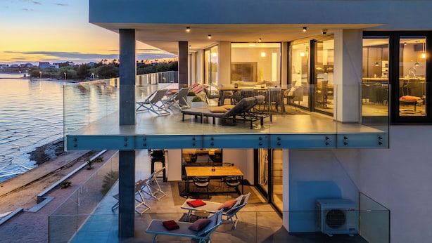 A modern building with a glass balcony overlooking a body of water, featuring outdoor seating and a dining area. - Jimmys Beach Privlaka: Penthouse dream, right by the sea and heated pool (Privlaka)