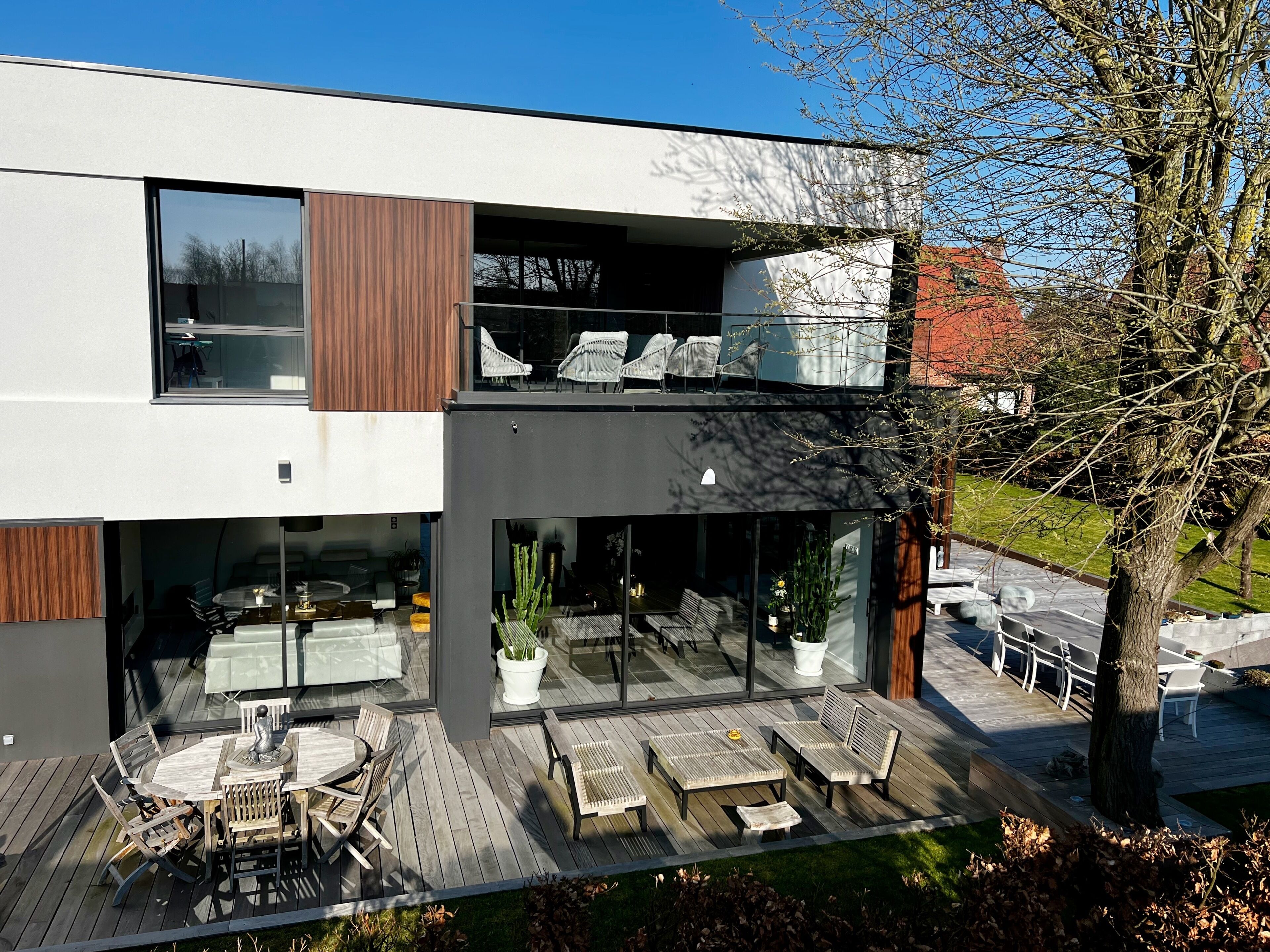 Una casa moderna de dos pisos con terraza, mobiliario exterior y ventanas grandes.