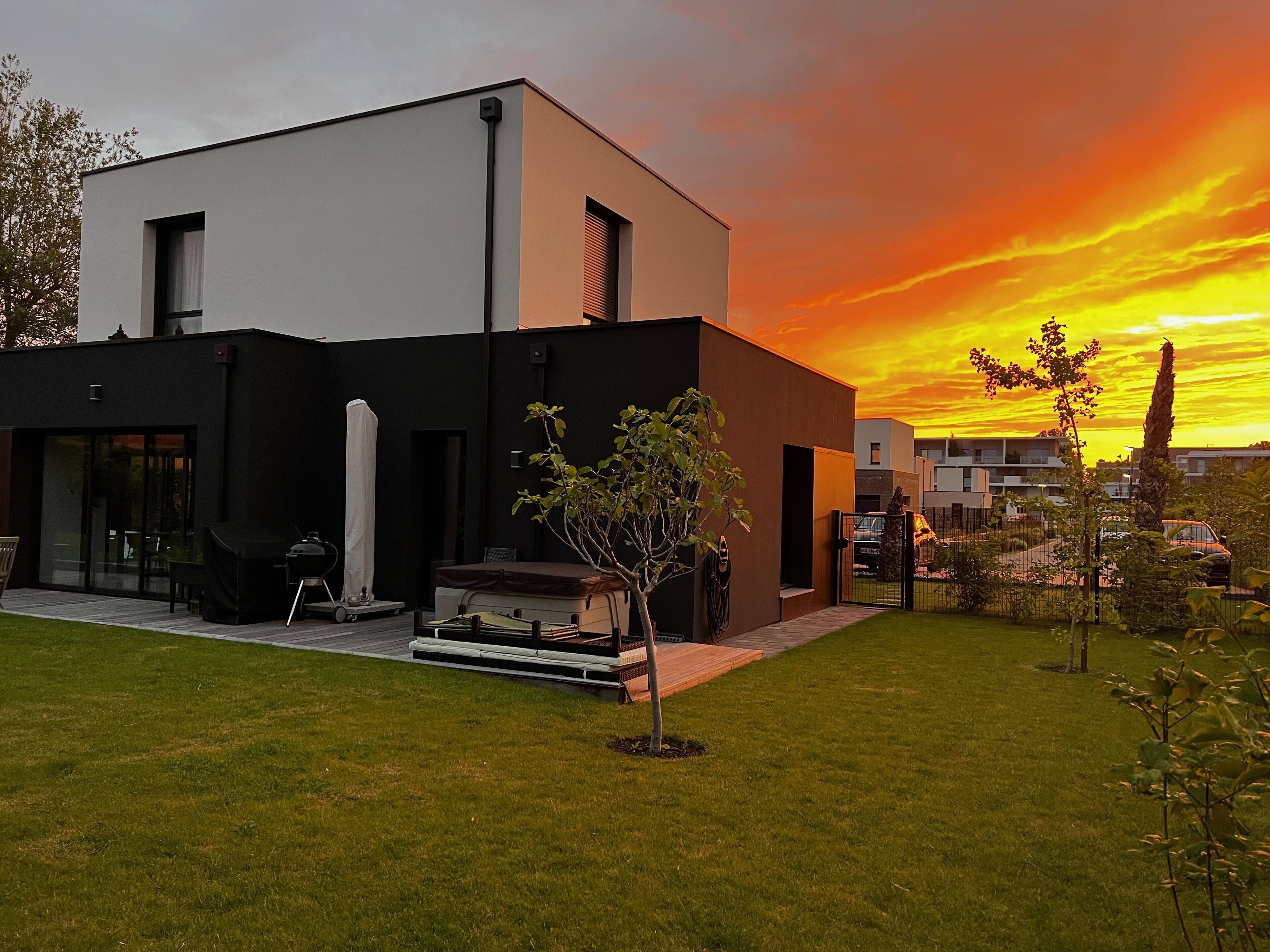 A modern two-story house with a flat roof, large windows, and a small front yard with a tree and a barbecue grill.