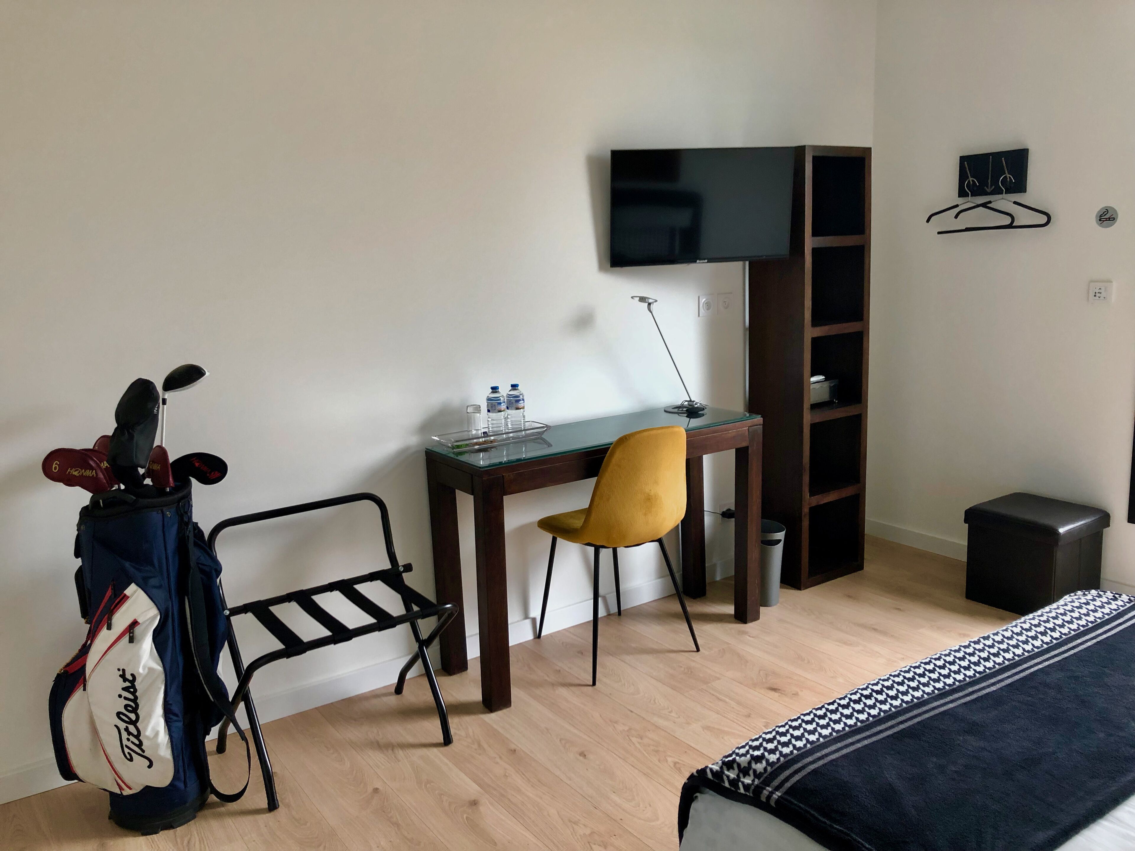 A hotel room with a bed, a desk with a chair, a TV, a coat rack, and a golf bag.