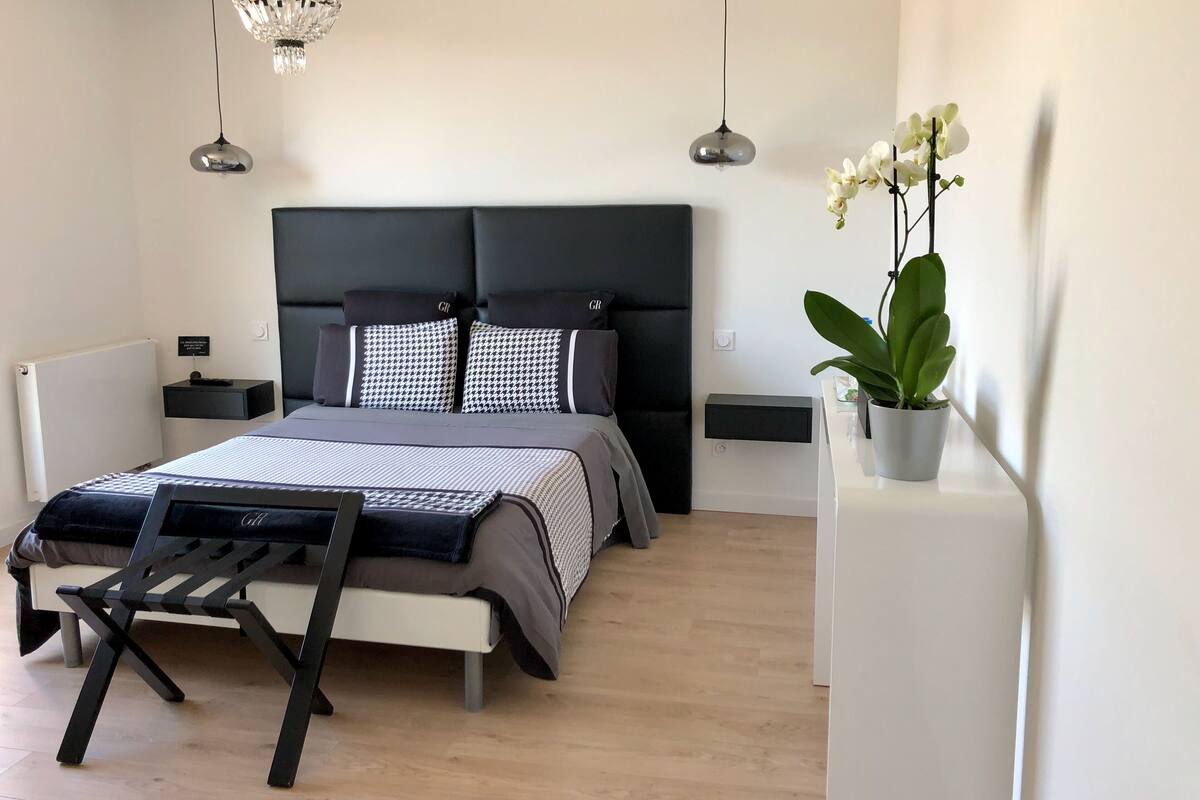 A modern bedroom with a bed, bedside tables, a nightstand, a hanging light fixture, and a potted plant.