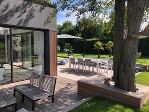 An outdoor patio with a glass door leading to a modern interior, a wooden deck, and a grassy lawn with a tree and a barbecue area. - Hôtelya - Lille - Villeneuve d'Ascq (Villeneuve-d'Ascq)