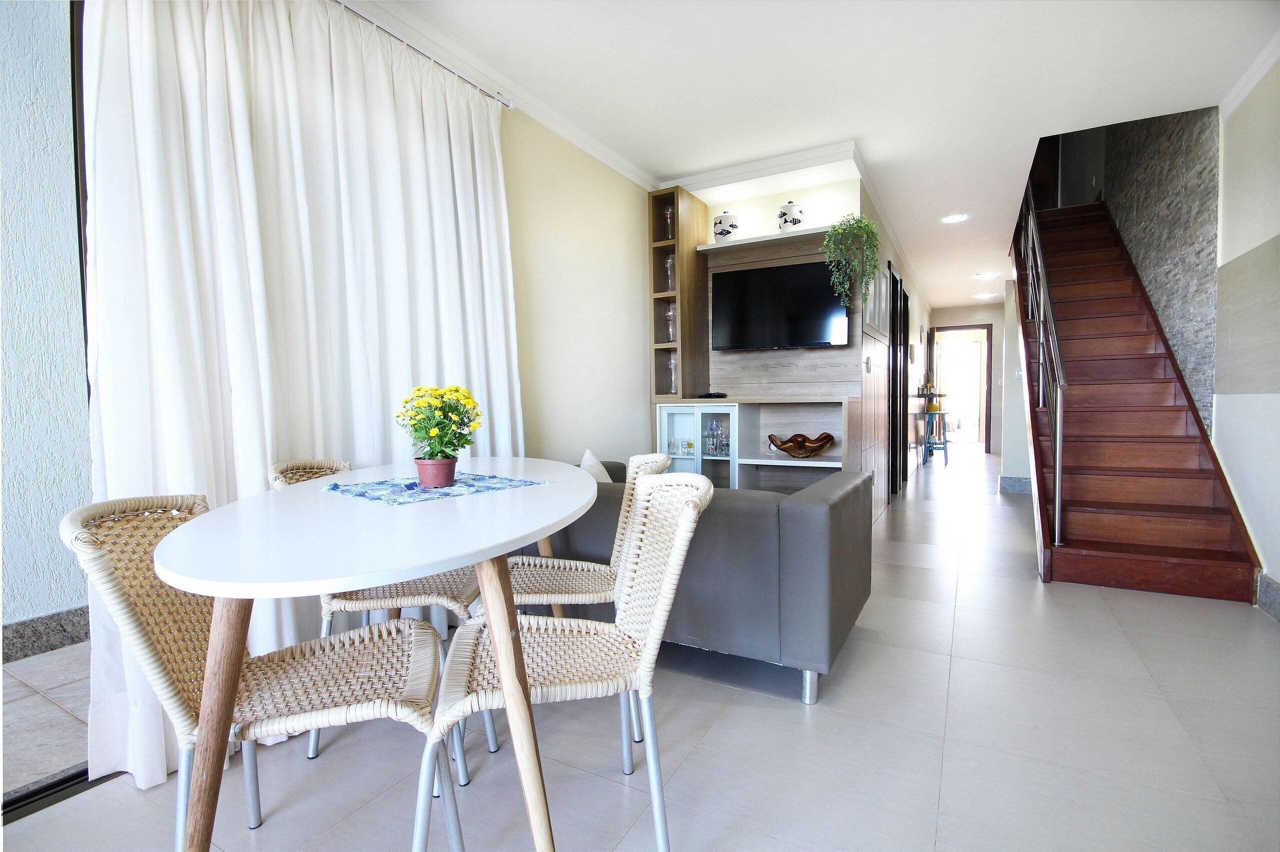 A modern living room with a round table, wicker chairs, a grey sofa, a TV mounted on the wall, and a staircase.