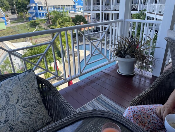 A balcony with wicker furniture, a potted plant, and a view of a pool and buildings.