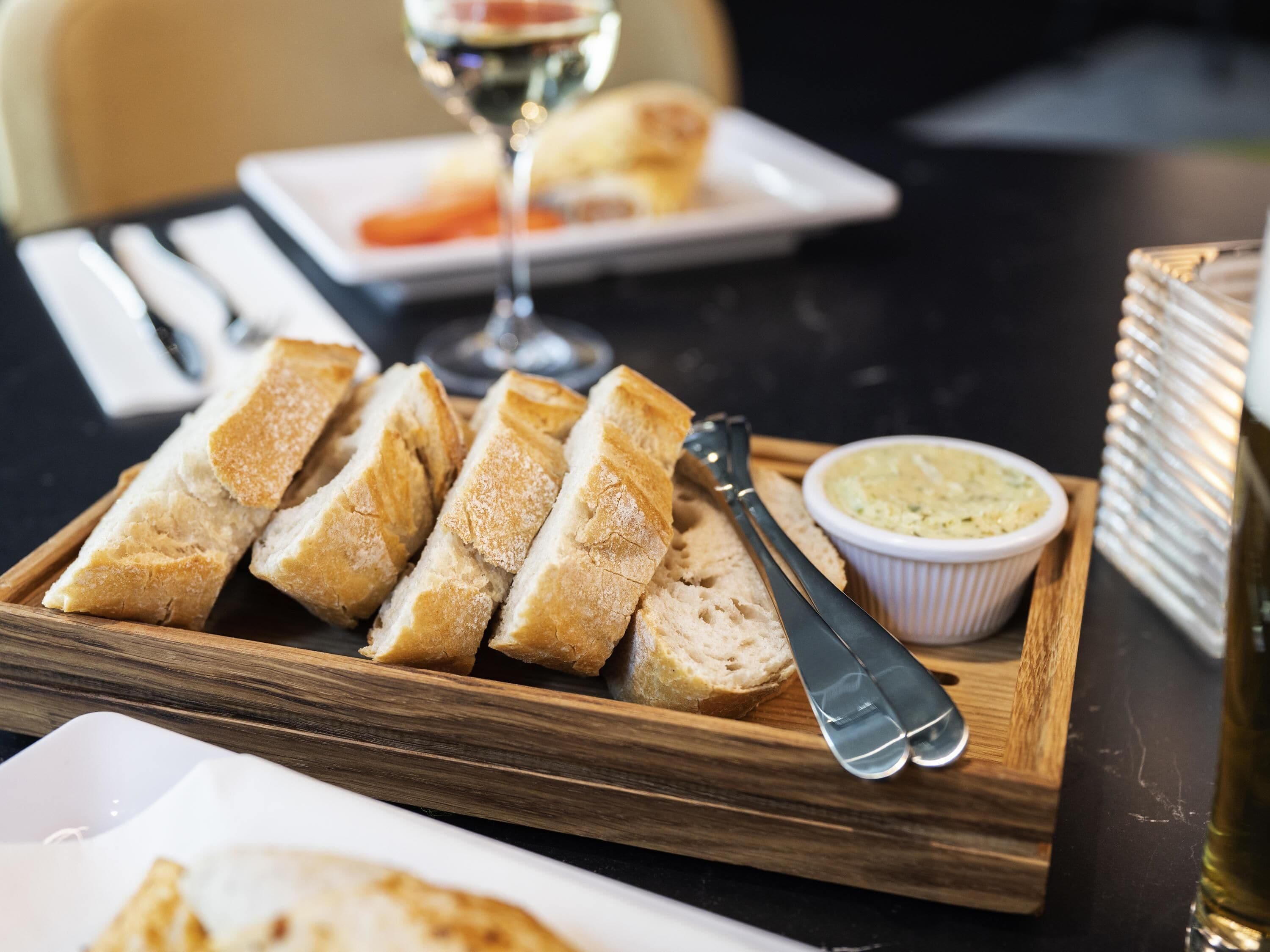 A wooden serving tray with sliced bread and a small bowl of butter on a table.