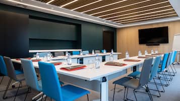 A conference room with white tables, blue chairs, and a large flat-screen TV on the wall.
