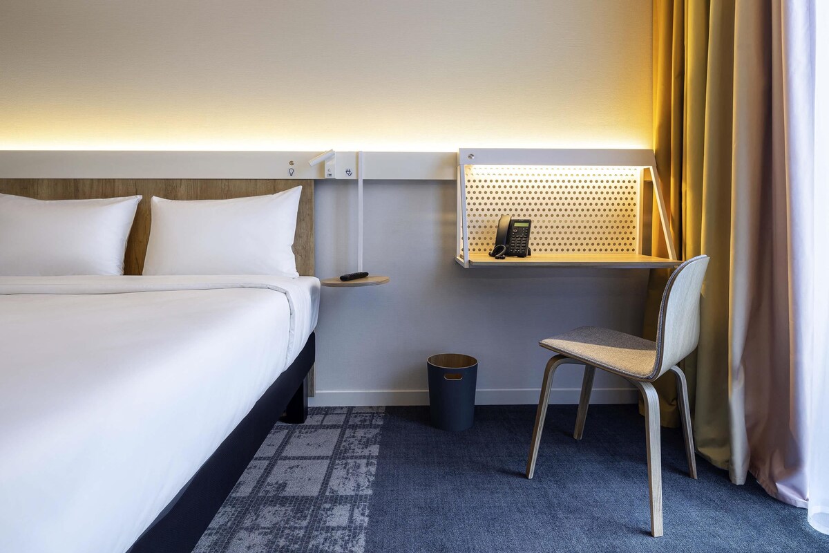 A hotel room with a bed, a desk with a phone, a chair, a waste bin, and a wall-mounted shelf.