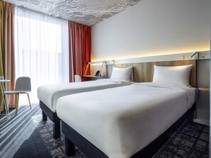 Ein Hotelzimmer mit zwei Betten, einem Schreibtisch, einem Stuhl und einem großen Fenster mit Vorhängen.