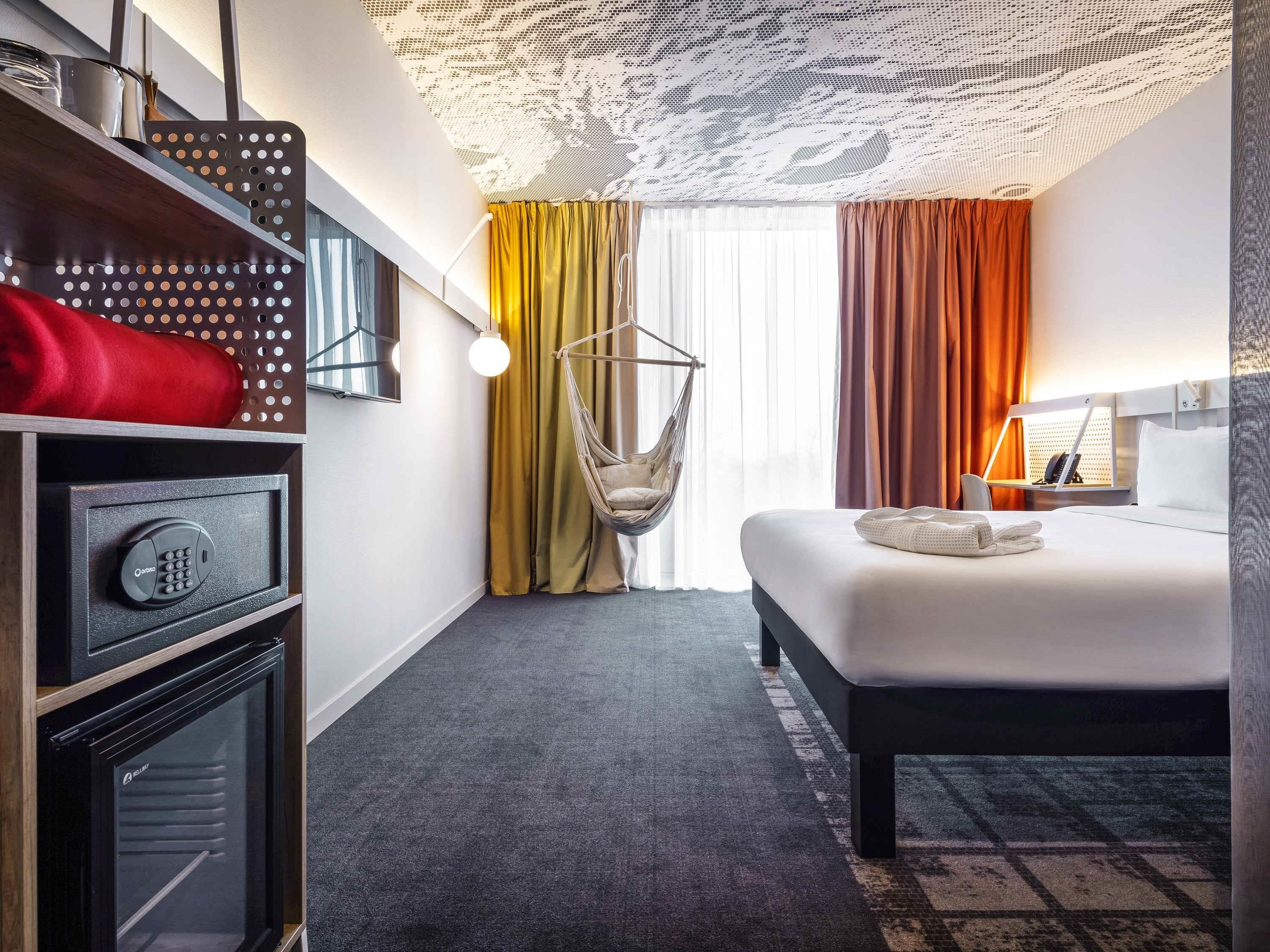 Una camera d'albergo con un letto, una sedia a dondolo, una cassaforte, un minibar e un'ampia finestra con tende.