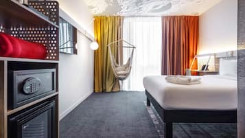 Una camera d'albergo con un letto, una sedia a dondolo, una cassaforte, un minibar e un'ampia finestra con tende.