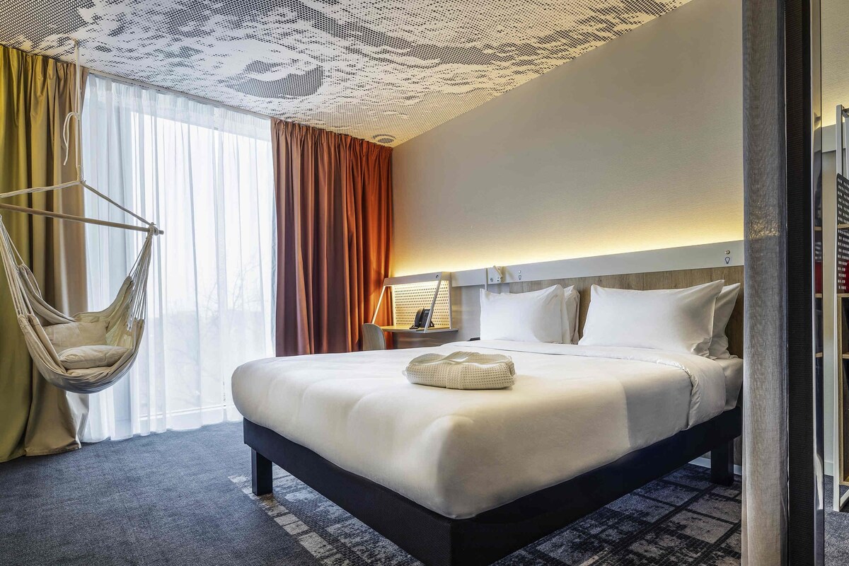 A hotel room with a large bed, a hanging chair, a nightstand, and a lamp.