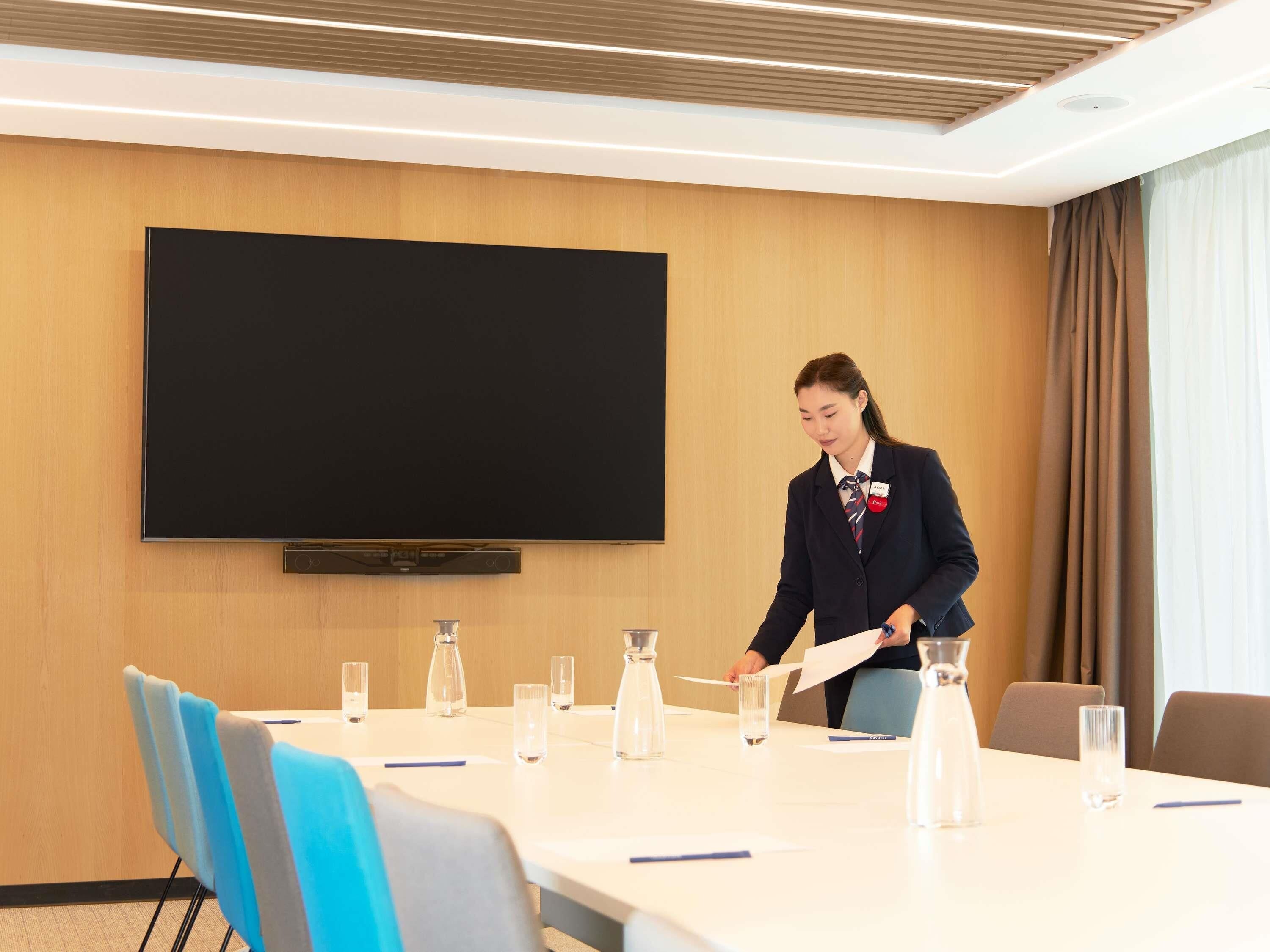 A conference room with a large flat-screen TV, a long table with chairs, and a person standing near the TV.
