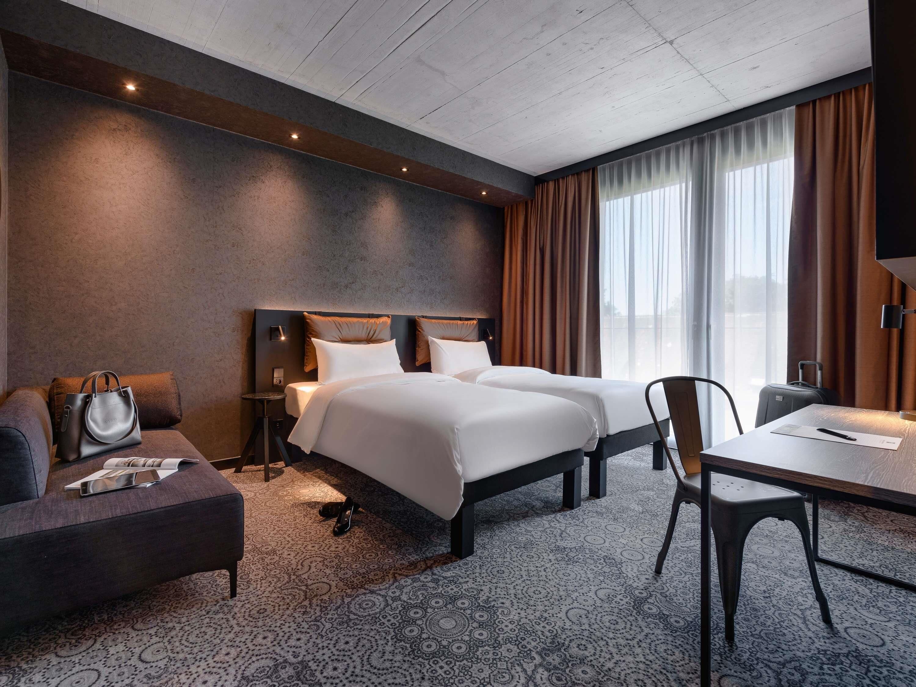 A hotel room with two beds, a desk, a chair, a sofa, and a handbag.