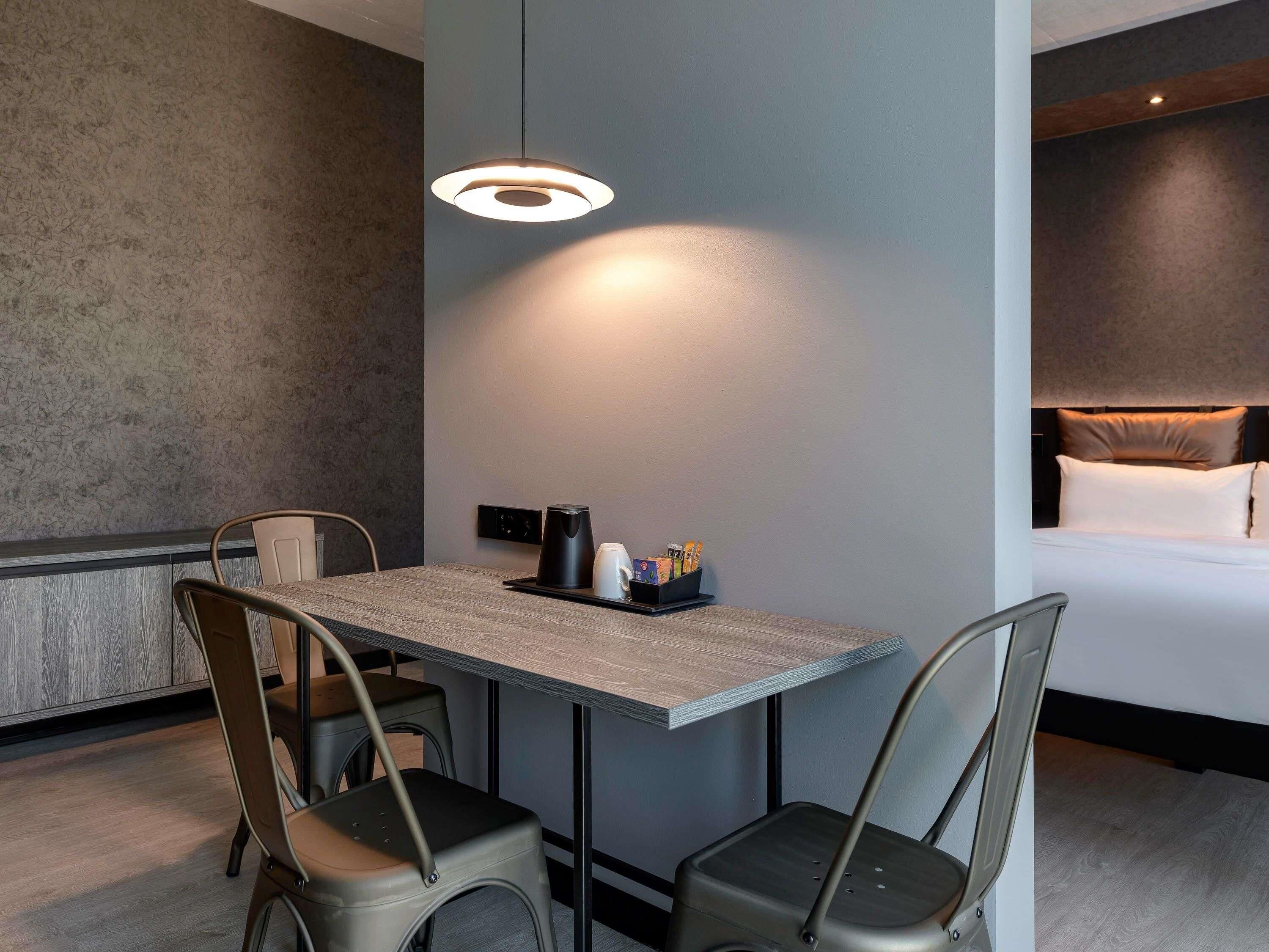 A modern hotel room with a dining area, a bed with a headboard, and a coffee maker on the table.