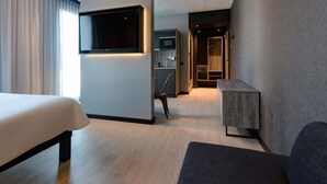Ein modernes Hotelzimmer mit Bett, Wandfernseher, einer kleinen Sitzecke und einer Küchenzeile im Hintergrund.