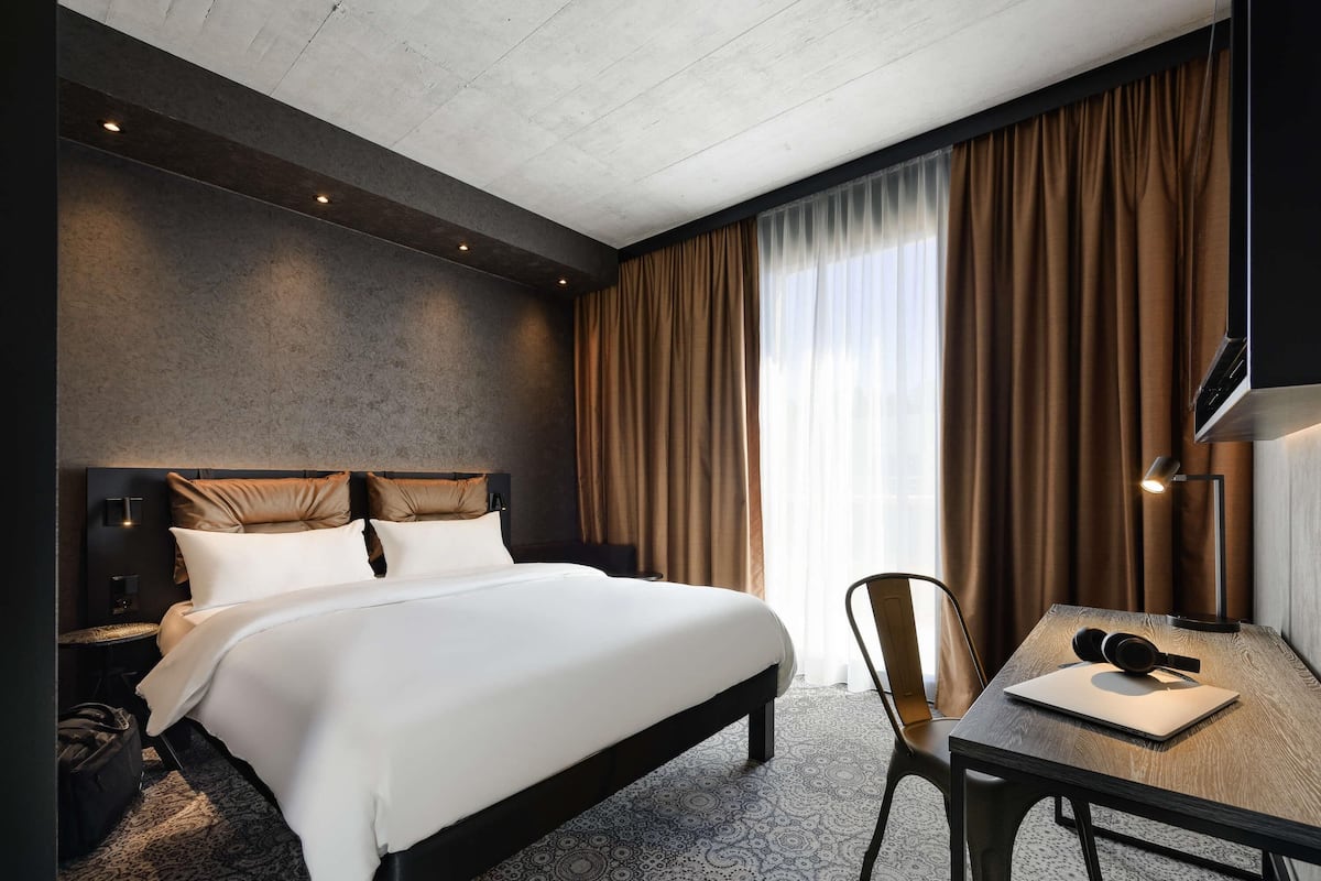 Une chambre d’hôtel avec un lit, un bureau avec un ordinateur portable, une chaise, une télévision et une fenêtre avec des rideaux.