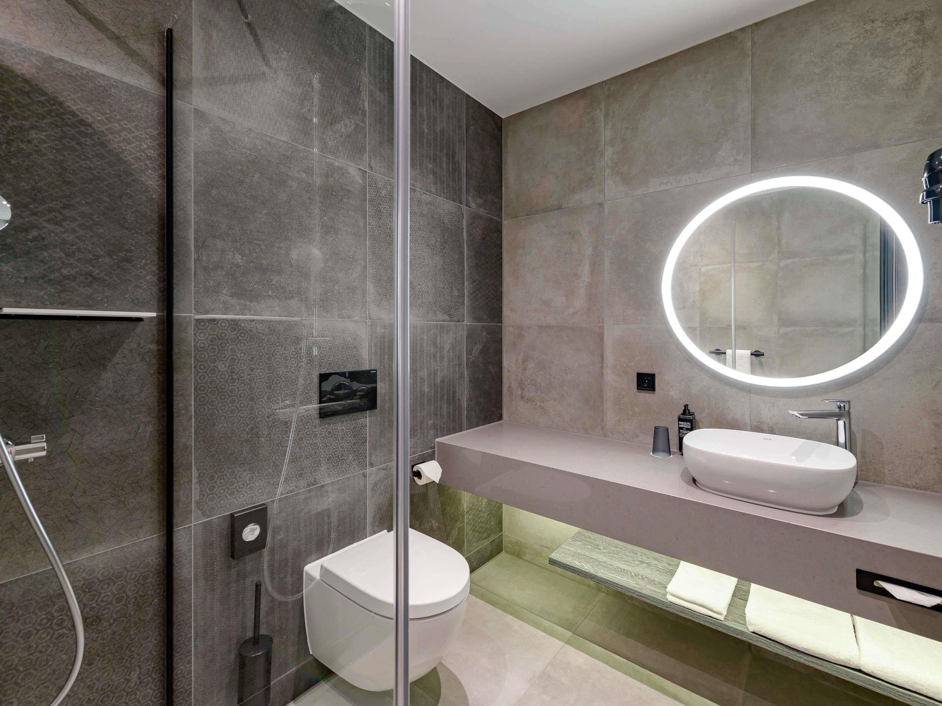 A modern bathroom with a glass-enclosed shower, a round mirror with a light, and a white sink.