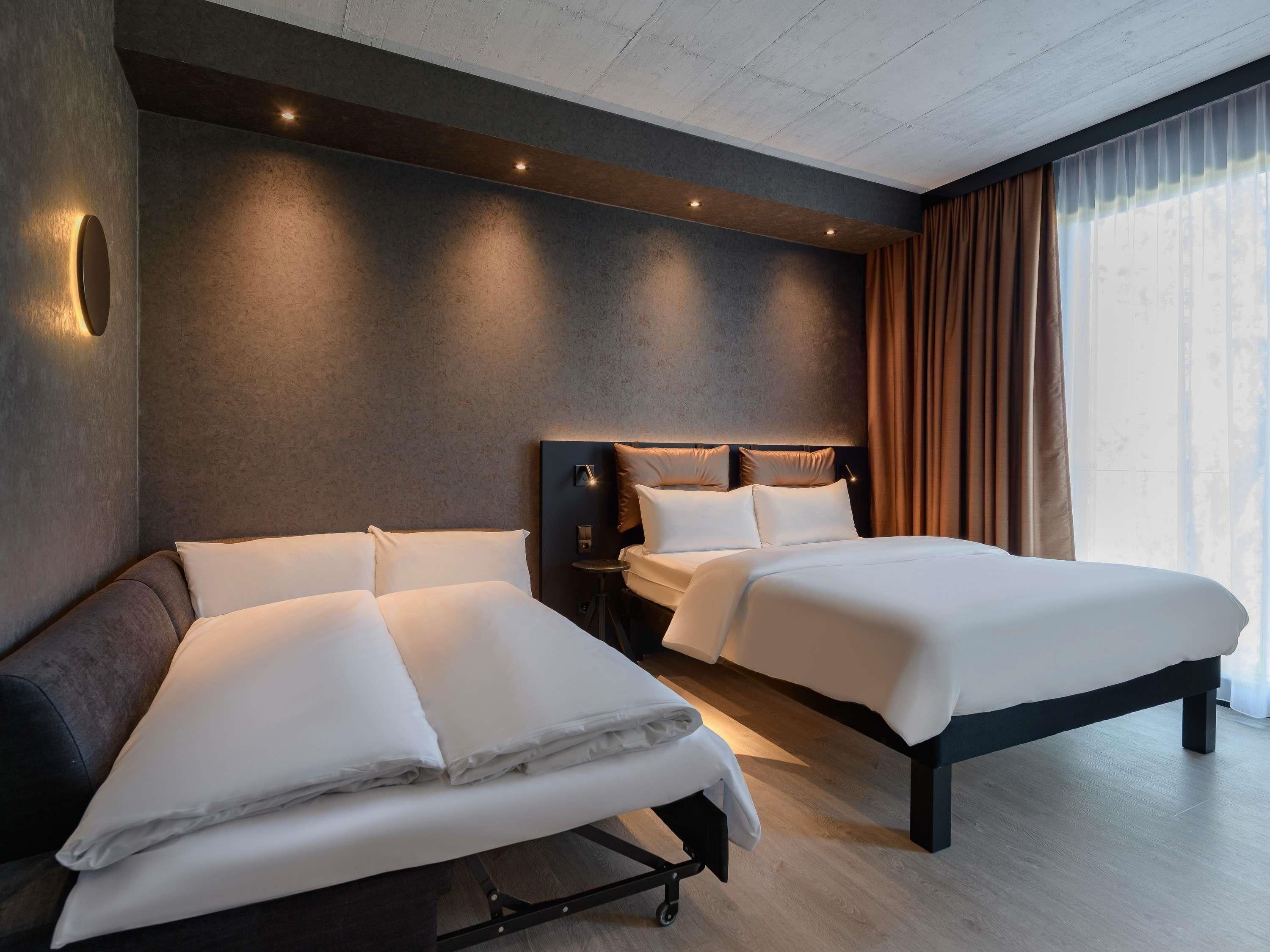 Ein modernes Hotelzimmer mit zwei Betten, einem dunklen Kopfteil und einem großen Fenster mit Vorhängen.