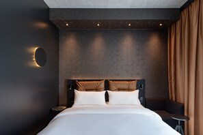 Une chambre d’hôtel moderne avec un grand lit, deux oreillers, une table de chevet, une petite table et un luminaire fixé au mur.