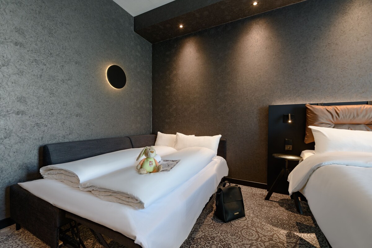 Une chambre d’hôtel moderne avec un grand lit, un plus petit lit, une applique murale ronde, une petite table et une valise.