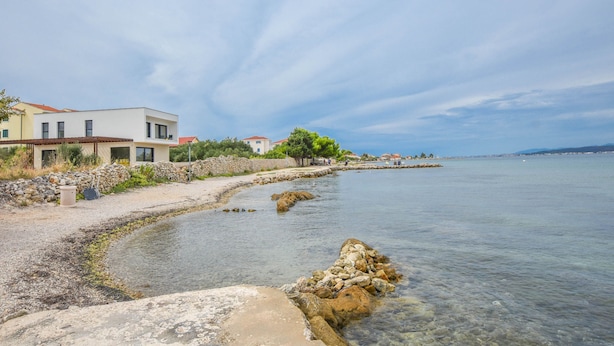 A coastal property with a stone wall, a clear view of the sea, and a modern building with a red roof. - Apartments Taya (Dobropoljana)