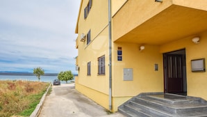A yellow building with a staircase leading to a dark door, a small tree, and a car parked in front. - Apartments Taya (Dobropoljana)