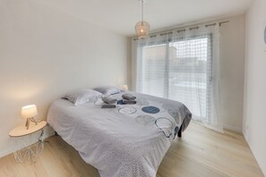 Et soveværelse med seng, sengetøj, et vindue med lette gardiner og en hængende lampe.