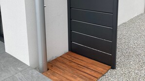 A modern gate with horizontal slats, a metal pole, and a wooden step leading to a gravel path. - La Roskoker - Villa 4 chambres et piscine privée proche plage - Roscoff (Roscoff)