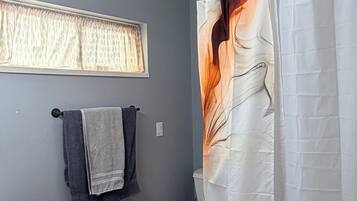 Une salle de bain comprenant des toilettes, un lavabo, une fenêtre avec des rideaux et un rideau de douche orné d’un motif de feuilles.
