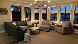 Una sala de estar con sofá, sillones, una mesa de centro y una mesita auxiliar con lámpara. Amplias ventanas ofrecen vistas a un cuerpo de agua y el horizonte de una ciudad.