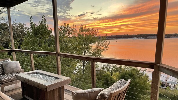 Terraza de madera con mobiliario exterior, incluyendo un sofá y sillas, con vista a un cuerpo de agua durante el atardecer.