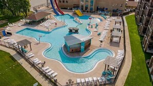 Una piscina con tobogán, tumbonas y un pequeño edificio, rodeada de césped y un edificio al fondo.