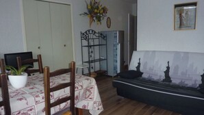 A dining area with a table set for two, a sofa with a cityscape print, a potted plant, and a metal shelf unit. - T3 PRA LOUP 56 M2 Magnificent valley and mountain views 2 bedrooms (Uvernet-Fours)