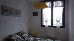 A bedroom with a bed, a window with sheer curtains, a yellow hanging light, and a small bedside table with a lamp. - T3 PRA LOUP 56 M2 Magnificent valley and mountain views 2 bedrooms (Uvernet-Fours)