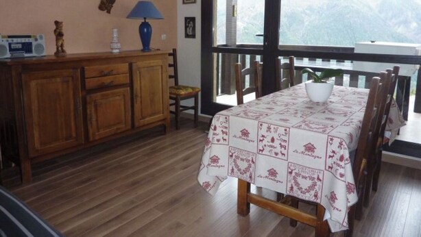 A dining area with a table set for two, a wooden cabinet with a radio, and a view of a mountainous landscape through large windows. - T3 PRA LOUP 56 M2 Magnificent valley and mountain views 2 bedrooms (Uvernet-Fours)