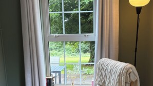Een gezellige kamer met een schommelstoel, een klein tafeltje met een boek, een vloerlamp en een raam met uitzicht op groen.