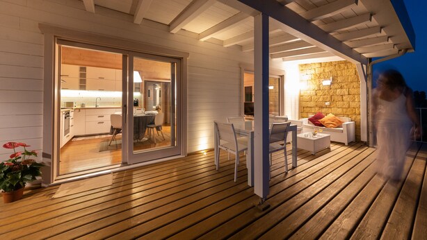 Um deck de madeira com porta de vidro que dá para uma cozinha e sala de jantar, uma parede de pedra e uma área de estar externa com mobiliário branco.