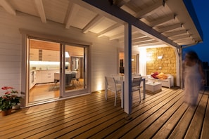A wooden deck with a glass door leading to a kitchen and dining area, a stone wall, and a white outdoor seating area.