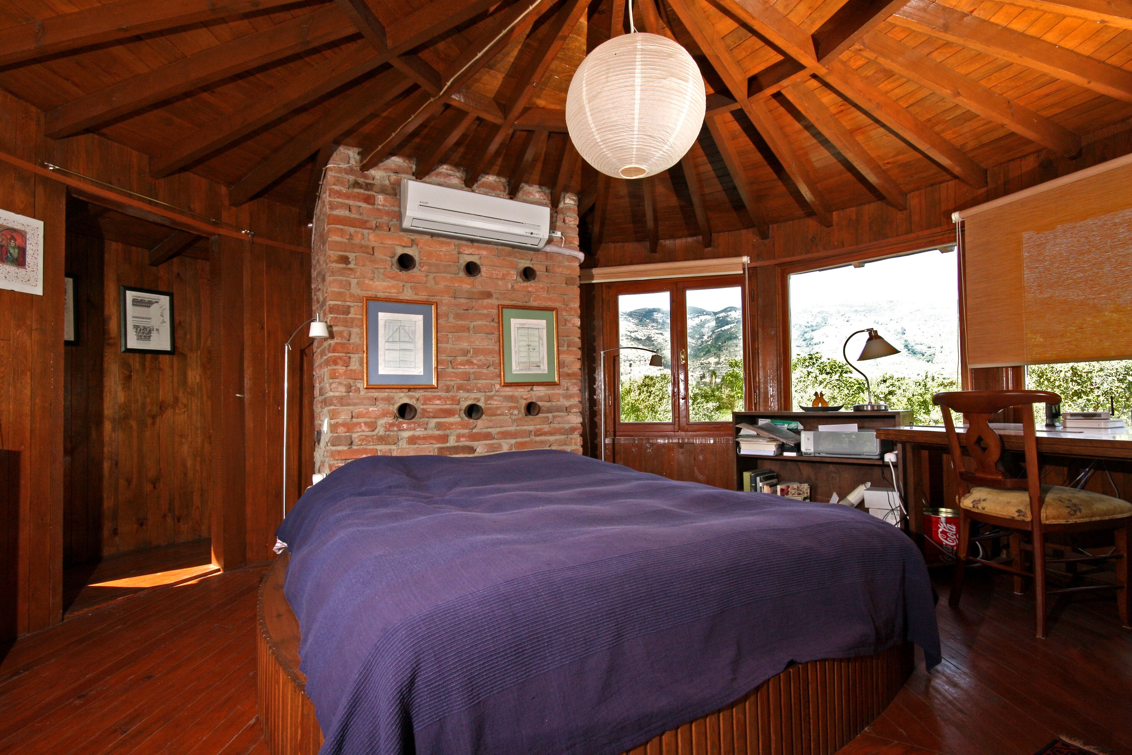 A rustic wooden room with a bed, a desk with a chair, and a large window.