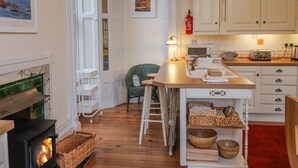 Een gezellige keuken met een open haard, houten vloeren, witte kastjes en een eethoek met rieten stoelen.